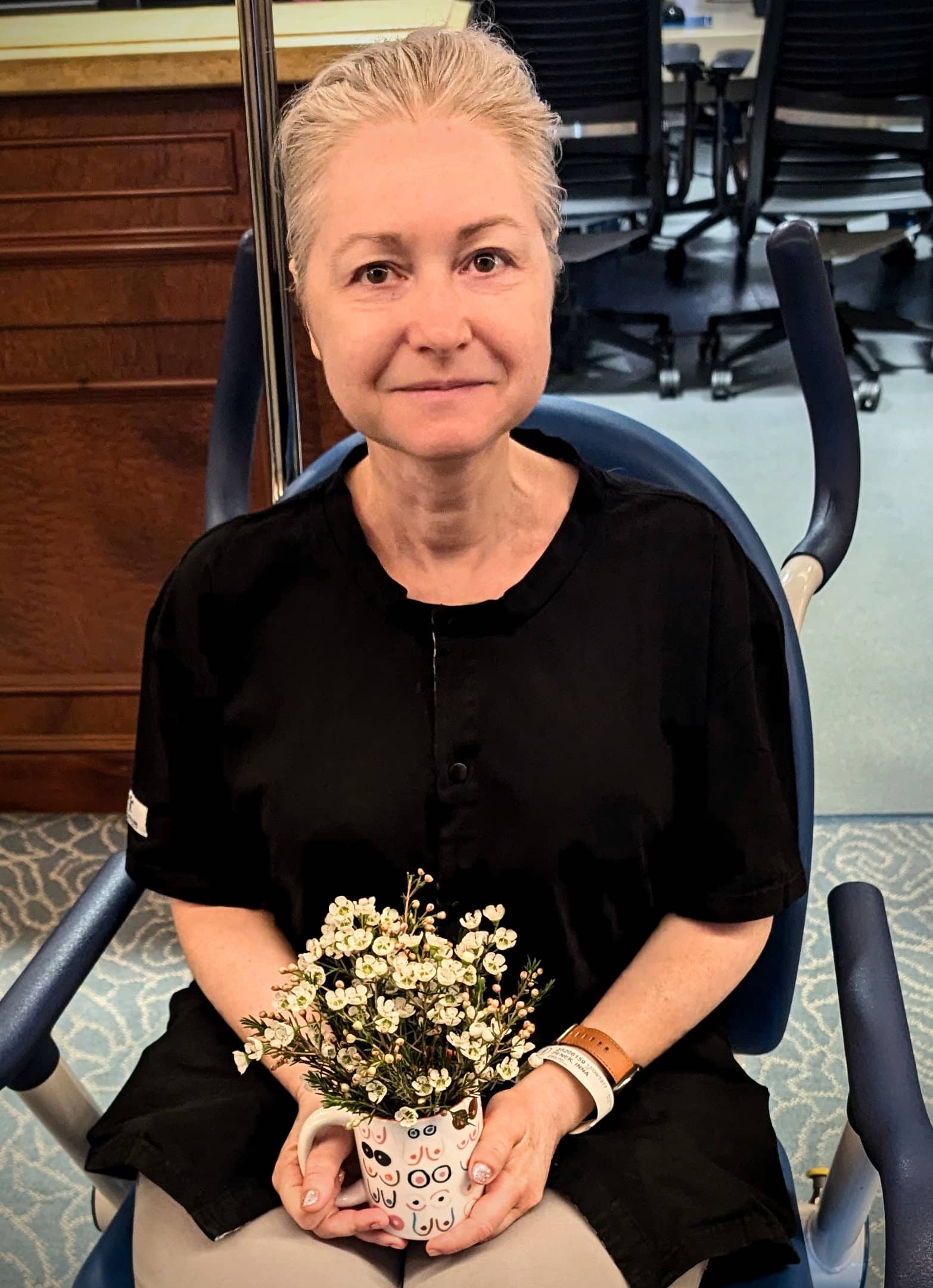 A woman with short gray hair sits in a chair, wearing a black shirt and holding a mug filled with small white flowers—perfect for inspiring bat mitzvah project ideas. Office chairs and desks are visible in the background.