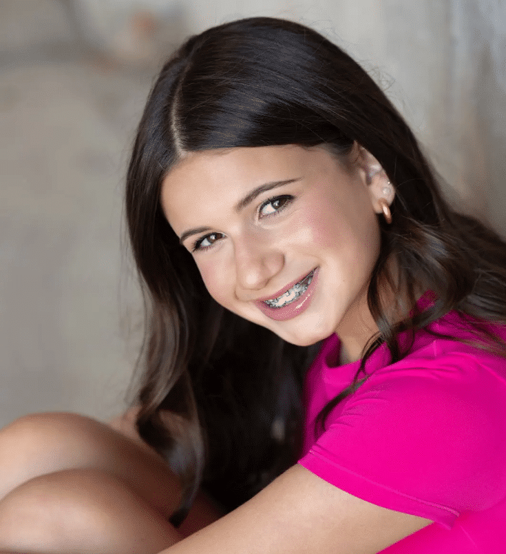 A girl with long dark hair, wearing a bright pink top and braces, sits with her knees up and smiles at the camera against a blurred neutral background, perhaps brainstorming bat mitzvah project ideas.