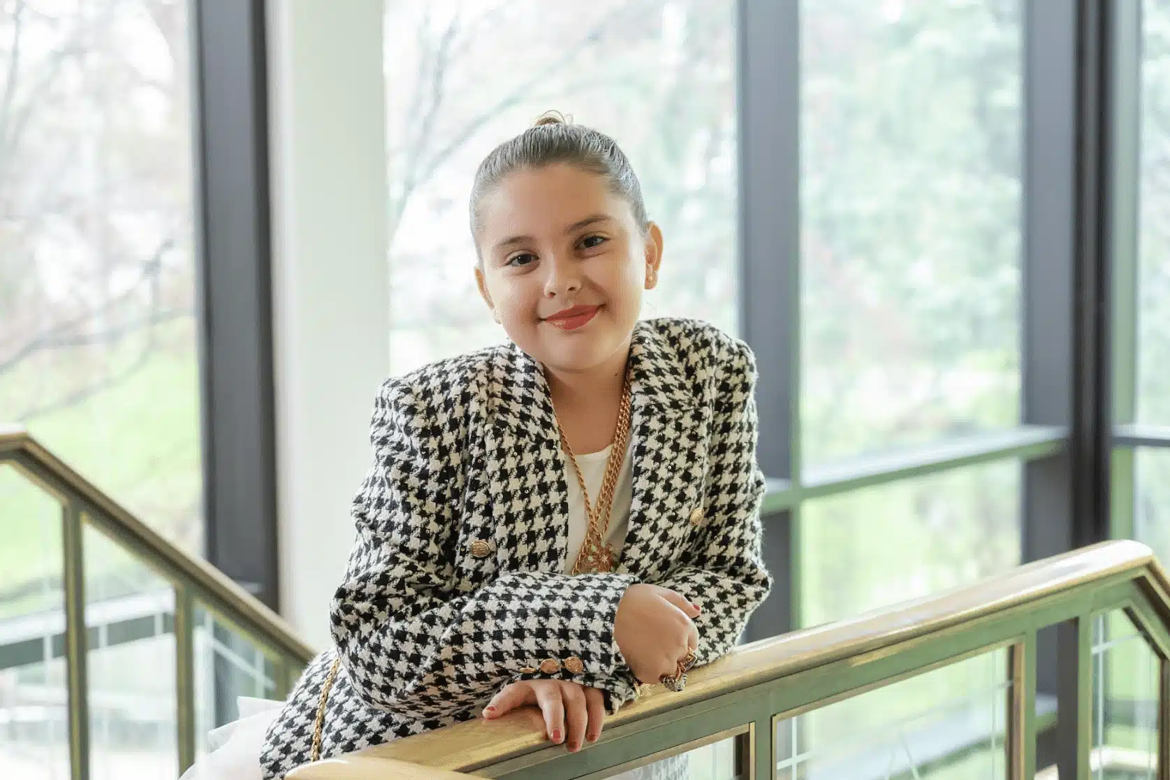 A young girl with her hair tied back, wearing a black and white houndstooth jacket, leans on a railing indoors, smiling at the camera. Large windows and greenery are visible in the background, perfect inspiration for bat mitzvah project ideas.