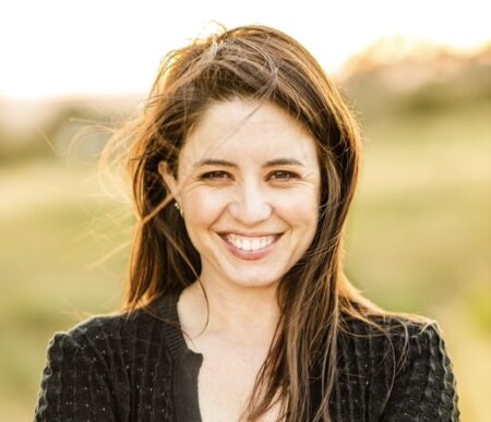 A woman with long brown hair smiles at the camera outdoors, wearing a black textured sweater. The sunlit, blurry background adds warmth, capturing the spirit of sharsheret stories in an uplifting outdoor moment.