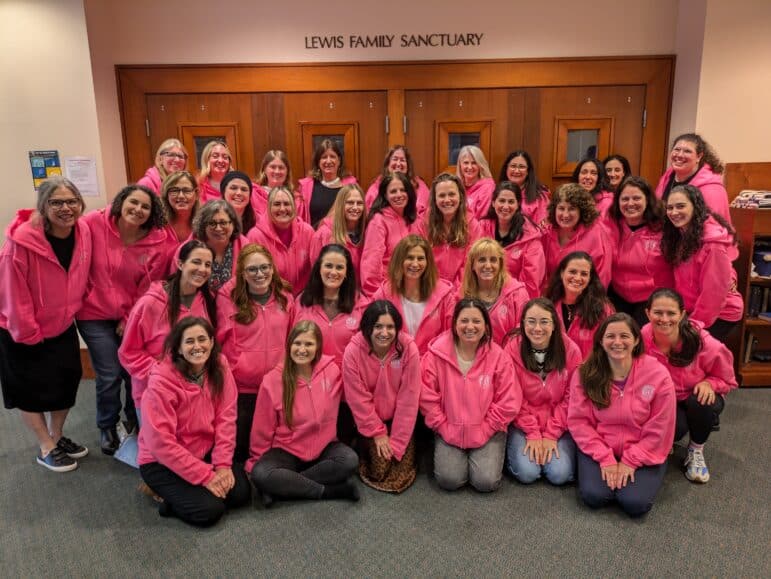 A group of about 40 women pose indoors, all wearing matching bright pink hoodies. They stand and kneel in several rows in front of wooden doors labeled “Lewis Family Sanctuary.”.